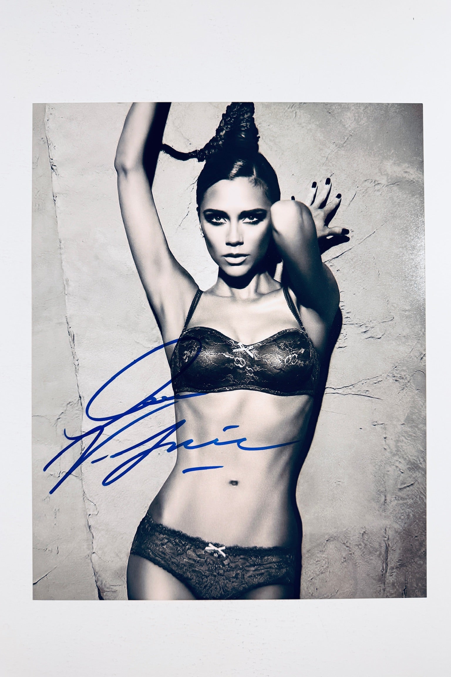 A black and white studio portrait of Victoria Beckham posing in lace underwear against a textured wall with her arms raised, featuring a blue marker signature across the left side.