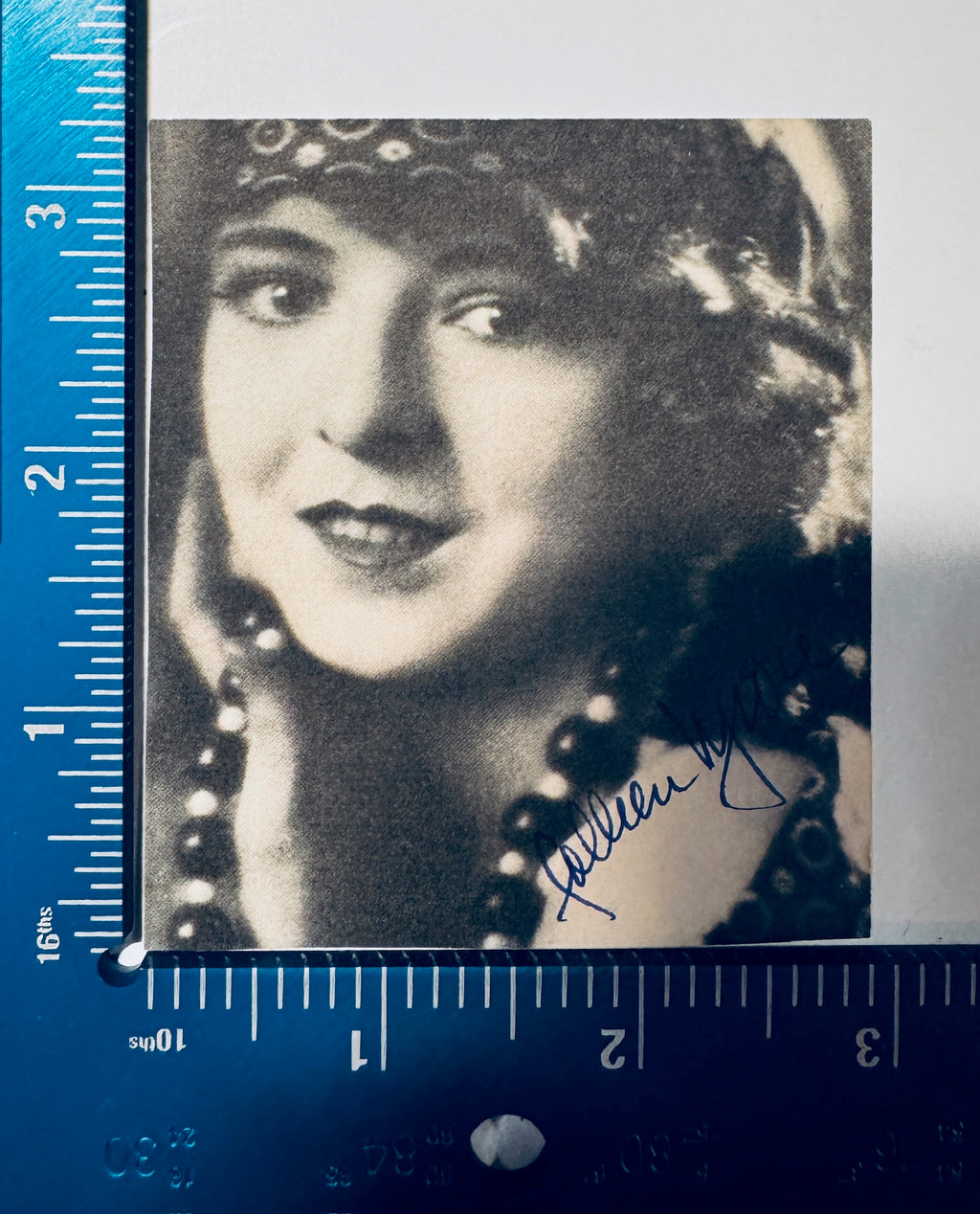 Vintage sepia-toned movie star magazine clipping headshot of Colleen Moore wearing a pearl necklace, aligned with a blue plastic measurement ruler for archiving size verification.