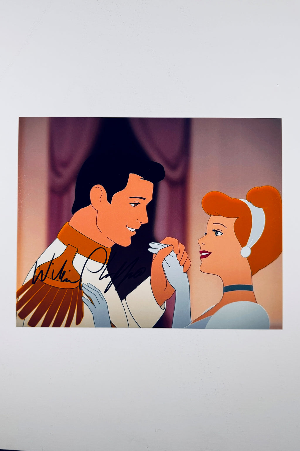 A vintage animation cel-style portrait of Prince Charming holding Cinderella's hand lovingly indoors against a purple curtain backdrop, featuring a black marker signature across the prince's shoulder and sleeve.