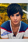 Signed 8x10 matte portrait photo of Zac Efron wearing a blue and white striped shirt with a necklace against an outdoor yellow foliage background, featuring a blue ink autograph across the lower-left area.