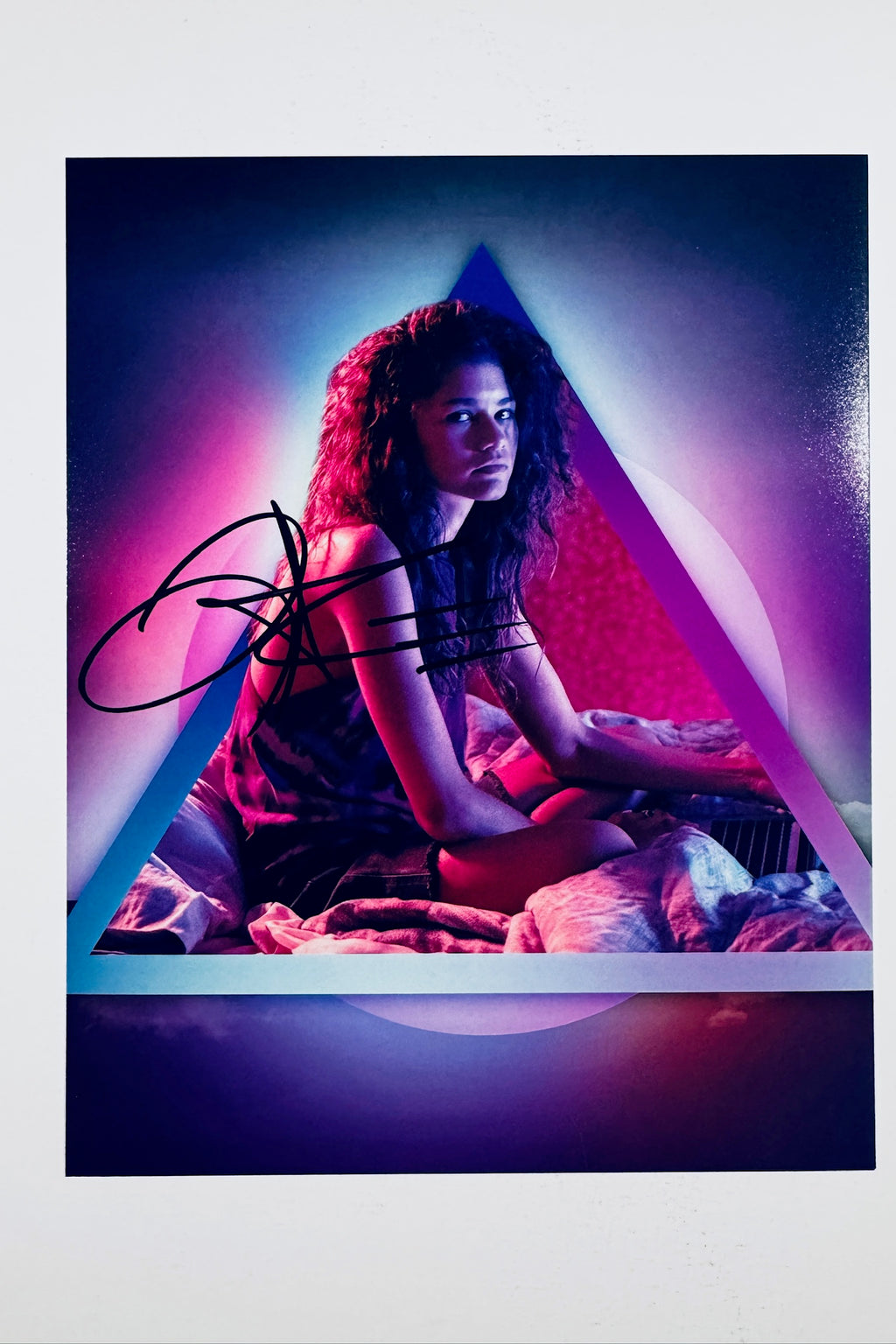 A portrait of Zendaya sitting on a bed framed within a glowing neon triangle and circle backdrop, featuring a black marker signature across the left side.